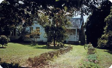 A white single storied house is set in a garden of mainly lawn and shrubs. The house is partly hidden by trees but a porch can be seen on the left with at least two gabled rooms to its left. The house is of Picturesque Gothic Revival Style with white stuccoed walls  and the roof is of shingles. 