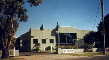 A plain rectangular building with a wide glass area marking the entrance. There are three square shaped windows on the left and the right wall is masked by trees but presumably has similar windows.The walls are rendered in a fawn colour. There are three flag poles between the side driveway and front lawn. One flies the Australian flag and the other is not easily identifiable. There appears to be a building at the end of the driveway but tree foliage is hiding all but a roof. There is a low front fence with two signs - one advertises a Tabaret and the other a Club Bistro. A large sign on the front lawn also advertises the Tabaret. A garden bed lies behind the fence. A corner of the adjacent property has a low white picket fence and there is a mature Eucalypt tree  on the road verge.