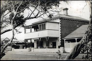 This photo shows a brick and stone 2 storey building. It has a wooden verandah to both floors with the upper floor enclosed in at one end. There is a small brick wall along the front of the property. There is a ti tree in the foreground. The roof is slate, and one chimney can be seen.