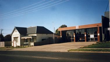 Front of a single storied building in the standard style of Victorian Fire Stations of the late 1900s. The letters CFA  and its logo are fixed on the decking above the central door.The building is set well back from the gutter behind a concrete apron. It has three vehicle bays behind glazed doors. A red fire truck stands in the middle bay with the door open. There is a single storied house to the left of the station. It’s walls are painted white and the pitched roof appears to be of grey corrugated iron. The front door is centrally placed off a porch which covers the right half of the facade. There is a “For Lease” sign on the small grassed front yard.