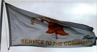 A flag is flying on a pole. It has a pale grey background with bronze coloured bell and lettering. The upper letters start Mornington and then a fold hides further letters. The lower line says “Service to the Community”.