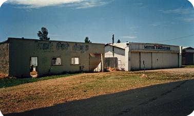 Two old single storied buildings rectangular in shape standing on open land on a slight slope. The building on the left is derelict while that on the right is in slightly better condition but appears to be neglected. It is coloured white with wooden doors occupying most of the facade. The architrave above has the words “MOTOR ENGINEER” painted on it.