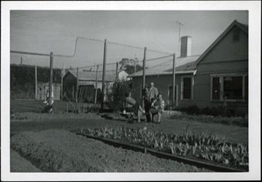 This photo shows the garden and tennis court of Tasma Court Hotel. There is a vegetable patch in the foreground. A group of people are in the garden, two women are seated, and a gentleman standing alongside. He is wearing a three piece suit, shirt and tie. There is a white weather board building to the right of the photo.
