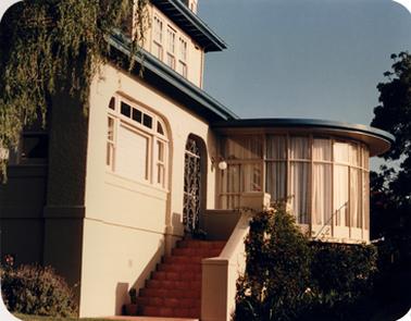 This is a photo of the front of a handsome ‘art deco’ house in cream rendered brick. A flight of nine or ten steps leads up to a front door with a decorated metal screen door. A main window to the left has a square of clear glass  central to surrounding small glazed windows in squares and rectangles. A second story is set back from the edge of the ground floor. A semicircle room extends forward on the right. It has a flat roof and is walled with curtained windows.A few shrubs frame the house.  