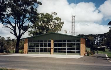 A large rectangular building in the style of contemporary fire stations. It is rectangular in shape with a low gable roof. The facade is dominated by two glazed doors flanked by narrow brick pillars. Presumed fire tankers can be dimly seen behind the doors. It stands on flat land beside a made road. A large tree stands to the left of the building with a line of pine trees to the rear. Two men are standing at the back corner on the right and there is a metal tower at the rear.