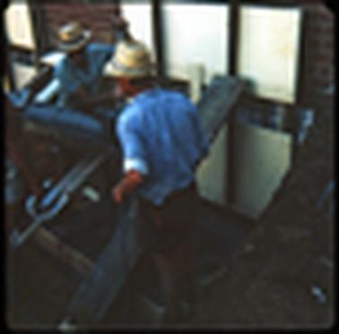 This photograph shows three work men working at the rear of a shop, smoothing down concrete. Two of the men are wearing blue shirts and white hats. 