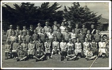 This photo shows 41 children and a teacher in arranged in four tiers. Some are in uniform, and some are in ordinary clothes. In the background are  several Cyprus trees. The teacher is a man. The boy in the centre of the front row is holding a sign stating “Mornington 2033 1959 Grande 5 & 6”.