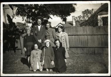 This shows a woman standing with six children around her in a garden. It appears to be winter as they are all wearing coats, and two are wearing berets. The three children in the front are kneeling. They are all smiling. 