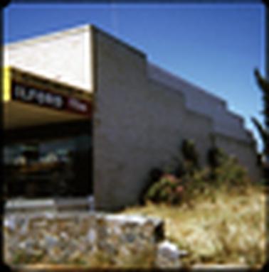 This image shows the side of a white brick shop. There is part of the sign visible on the shop front and it is the Chemist shop. There is part of an advert for film visible too. In the foreground is a small rock wall which is broken at the end, and on the left of the picture there is a white picket fence. 