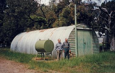 A Nissan hut stands in front of mature trees on grass beside a gravel track. It is covered in curved corrugated iron with a door on the end wall of weatherboard. A water tank on a stand is beside the hut along with two men in casual clothes.