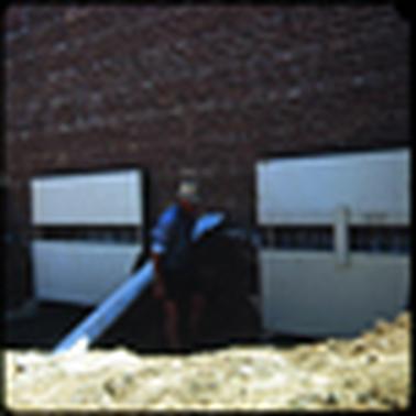 This photograph shows a work man wearing dark shorts, a blue shirt and a white hat. He is working against a red brick wall, which is the rear of a shop.