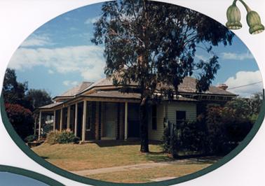 A dark brick house of a single story has a tiled double pitched roof and a simple verandah supported by simple white painted posts. The A tall corner window appears to be decorated with tuck pointing in cream brick. There is a small weather board room added at the right hand end of the verandah. The house sits on a simple, raised grassed area with several shrubs and a small eucalypt which is close to the street. The property is partially fenced on the right.