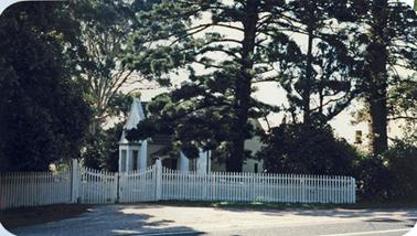 A small white Gothic revival house behind a low, white picket fence with closed double gates and a gravel entrance. It is surrounded by trees including a eucalypt, a fine specimen of a pine tree and shrubs. It stands beside a main road with an asphalted surface. 