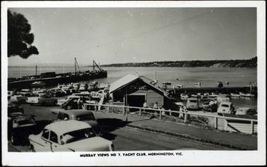 This photograph shows a weather board building on the foreshore, with the Pier to the left. There are many cars parked close by. There are a number of small boats moored alongside. People are shown around the club.