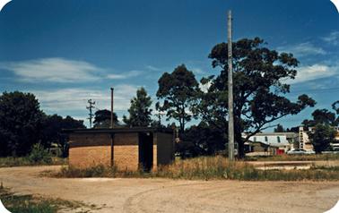 A gravel surfaced road curves around a small yellow brick building in the left foreground. There are mature trees on rough land between the building and a road in the background. There is a white building on that road which is a timber and hardware store.  