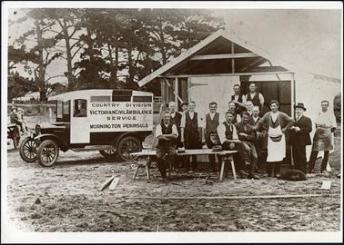 The image shows to the right of photo a group of adults, to the left of the photo is a c1920 ambulance, written on the side of the vehicle 'Country Division Victorian Civil Ambulance Service Mornington Peninsula'