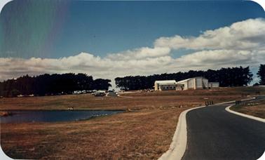 Long distance view of of a group of cream coloured buildings set in a wide expanse of sun browned grass. An asphalted road winds past a lake and leads to a car park containing several cars to the left of the buildings. A line of pine trees marks the horizon.