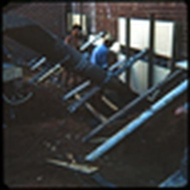 This image shows two work men pouring concrete at the base of a red brick wall. It is evidently the rear of a shop in Main Street. The older man is wearing a blue shirt and a white hat. The younger man is bare chested. 