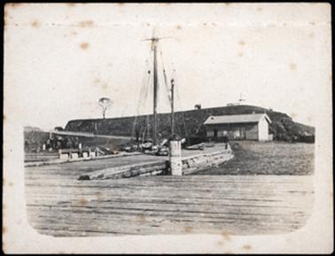 This is a photograph taken from the Pier toward the land. There is a shed type building where the Pier meets the foreshore. On the crest of the hill are a group of people. There is a boat with a tall mast on the far side of the Pier. In the centre of the image is a bollard.