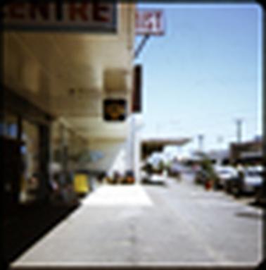 This is a view along Main Street of the shop fronts. In the foreground is a Chemist, and a furniture shop with some chairs outside is in the centre of the photo. The sky is blue and it looks like a warm summers day. There is a woman looking in a shop window, and there are many cars parked along Main Street.