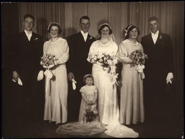 This photograph shows a wedding party. The group consists of a bride, groom, two bridesmaids, and a little girl maid of honour. The three women each have a head dress. The bride has a flowered tiara, and the bridesmaids have a plainer version. They have long gowns, and large bouquets. There are three men shown, the groom and two groomsmen. The men are carrying white gloves. The little maid of honour is holding a basket of flowers.