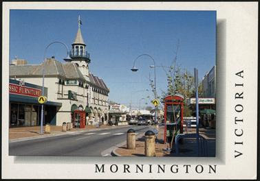 This is a view of Main Street showing the Grand Hotel. The view is looking away from the beach. It is taken from the opposite side of the road to the hotel. There are two red telephone boxes shown, one on either side of the road. The Classic Furniture shop is adjacent left to the Grand Hotel and to the right a real estate office. There is a cloudless sky, bright blue.
