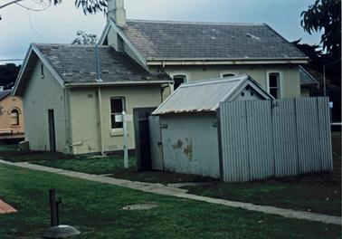 The photograph is taken from the rear and shows a substantial building of stone with a slate roof and a smaller shed of fibro cement with a metal roof. The main part of the building is double gabled  with a slate roof. The middle section is the larger and has three windows set in the side wall. The rear section is smaller with one window. on the side wall and a simple door at the rear. There is a chimney set in the roof at the rear of the middle section. The shed has corrugated iron fences on either side. There is a wooden door on the left. The buildings stand on flat lawn and there is a paved path at the rear of the buildings. Portion of a stone building can be seen across a road. 