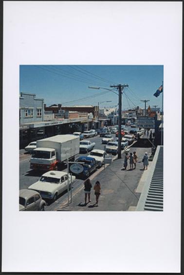 This is a view of Main Street from the top of a building looking down. It is a sunny day, and there are several people shown in summer clothes. There are lots of cars parked down either side of the street and traffic on the move. To the right of the image is a telegraph pole, and a real estate office. and the Australian flag can be seen at the top right of the photo.