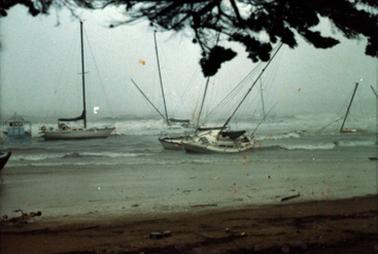 This shows some yachts damaged in a storm. The day is grey and overcast. The masts of the yachts are broken, and one is submerged. There is another yacht on it’s side. Port Philip Bay is rough with white caps on the waves.