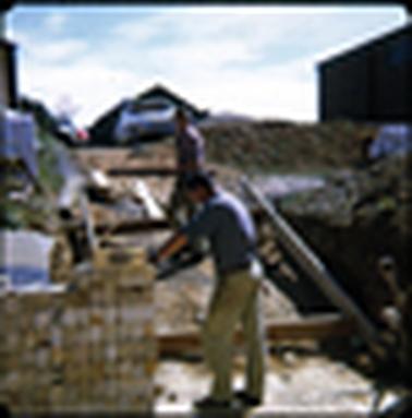 This photograph shows two work men working at the rear of the shops in Mornington. There is a white brick wall behind them with a ladder leaning against it. They are both inspecting a spirit level. 