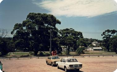 There is an area of land covered in gravel in the foreground. It is defined by a low post and rail barrier. A red collection bin near the barrier is next to a power pole  A wide grassed  verge containing mature trees lies between the barrier and a road. There are two cars parked on the gravel. 