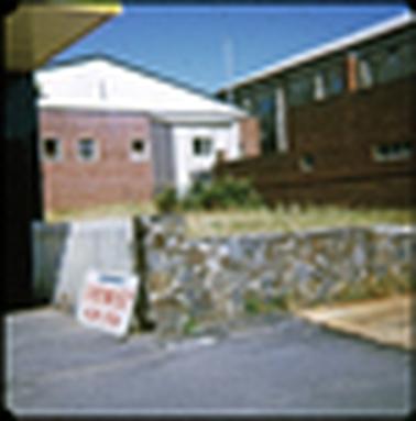 This photo shows the rear of a red brick industrial looking building. It has a pitched roof, and a small room added on. This small add on appears to be constructed off A.C sheeting with a tin roof. There are two small windows in the brick building. In the foreground is a low stone wall, which has a crazy paving look. To the left there is a picket fence, and a sign leaning against this “Chemist, now open”. 