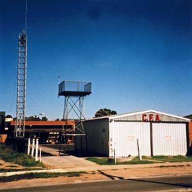 The rear of a terracotta coloured single story building is in the background with a tall metal tower on the left and a shed close to the footpath on the right. The shed has the letters CFA on its upper facade and beside it is a blue tank on a metal stand about double the height of the building and a pathway leads from the street to the rear building.
