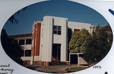 This is a two story building of part red brick and part white render. There is a substantial portico with steps in front and on the left side leading up to the white portico which has a set of four windows above the doorway. Brick steps appear to lead further up to the brick portion. There is an extension of the white surface to the right with CONVENT OF MERCY  on the upper facade. Behind the portico the left wing extends as a balconied two story structure. There is a circular driveway in front with several small trees and a small open fronted shelter is on the left. 