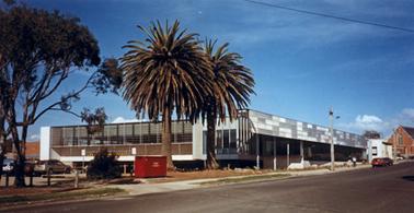 A large rectangular building is set on a corner block with  a car park beneath it, a gravel car park to its rear and a roadway to the right. A church is just in view opposite the corner of the building. The streetscape is bare of vegetation except for two palm trees, a eucalypt and a narrow strip of grass.