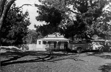 A more distant view of Glengala house 2 featuring a wooden picket fence and a utility truck parked in front of the fence. The house is painted weather board with a window on each side of the main door. On the left is a small enclosed sleepout with a door on to the front verandah. It has a skillion roof of corrugated iron.