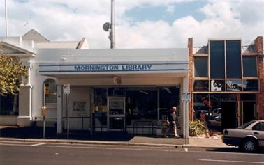 Single storied shop front between a restored older building and a modern glass fronted double storied shop/office building. A rectangular canopy carries the sign MORNINGTON LIBRARY. A pedestrian walks in front of the building. The footpath contains signs for Car Parking and a Bus stop. 