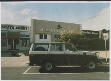 Image of Mt Martha Motors garage, brick construction, painted beige with 2 white painted double doors and 3 petrol bowsers. Four wheel drive parked on the road out the front. Bushes partly covering the right hand side of the building.