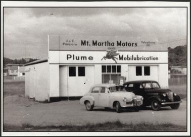 Image of a brick garage, painted white with two double doors. Two cars are parked in front of the building. The name Mt. Martha Motors is painted along the top of the garage. Domestic houses can be seen in the background. 