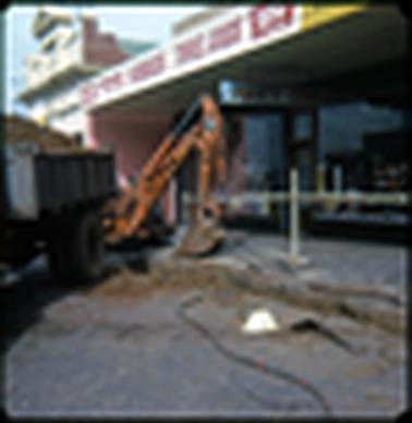 This shows the path outside a shop and some building work being done in the road, it appears to be concerning drainage. The shop has a sign across the front “Toys, Hobbies, Travel Goods”.  It also states “Variety Centre”. There is a digger shown to the left of the photo, and the rear of a truck can be seen. In the foreground there is a work man’s white helmet.