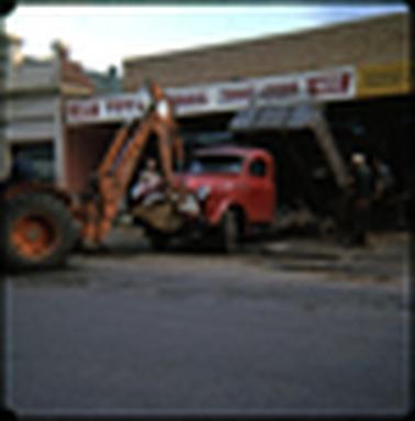 This shows the path outside a shop and some building work being done in the road, it appears to be concerning drainage. The shop has a sign across the front “Toys, Hobbies, Travel Goods”.  It also states “Variety Centre”. There is a digger shown to the left of the photo, and the front of a truck can be seen. The road is in the foreground.