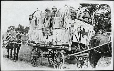 This shows a horse and cart, and riding in the cart are a group of young boys. The cart is decorated with hanging clothes, and walking sticks, and has a red cross on the front. On the left of the photo is another horse and cart, and the driver is wearing a helmet. 