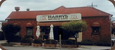 Red brick building with red corrugated iron roof and two large white umbrellas (closed) outside the front of the building. There are 4 plant boxes and two trees on the street and a large white sign over the front entrance.