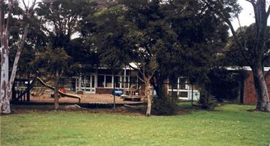 Timber and brick single storied preschool with windows along most of frontage. Photo shows building behind a high wire fence, and partially hidden by trees. Playground equipment visible behind wire fence. Photo title on back indicates land was owned by Downward family.