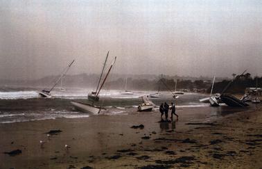 Stormy beach scene showing white water waves, and yachts being blown sideways,some wrecked boats on the beach, and clumps of seaweed washed up. Three people in a group on the beach appear to be looking at something one of them is holding. In the middle right of the photo is the shed with Boats for Hire painted on the roof.