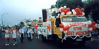 Main street scene with yellow truck decorated with Red Cross theme - red and white balloons, cloth banner with slogan “Laughter is the Best Medicine” and banner on side of a red cross on white background. A second white banner has a variety of hand-knitted socks , gloves and caps attached. Marching on the road beside the truck are boys dressed as doctors and girls dressed red and white as nurses. In the background are Main street shops, back from Queen Street.