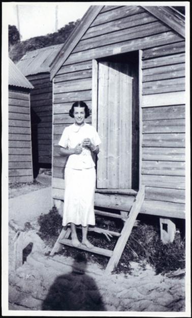 This shows a young woman standing on the steps of a bathing box. She is smiling and looks happy. She is wearing a two piece, skirt and blouse with a Peter Pan collar and short sleeves. Three bathing boxes can be seen in the photo. They are built of wood, with corrugated iron rooves.