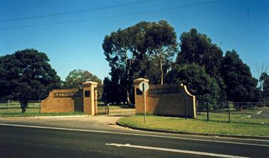 Cream brick walls flanking wrought iron gates, partly open to show gravel drive leading into park with large trees.