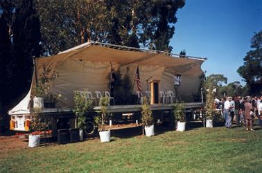 Temporary stage comprising a large flat tray (of a truck) on wheels, with a canvas awning erected as back wall and roof. Pot plants are on the stage and on the ground in front of the stage, and a microphone and lectern are set up centre stage. A group of well dressed people mostly wearing hats are standing to the right with backs to the photo. White plastic chairs and a large American and an Australian flag are on the stage, presumably at Balcombe Army Camp for 2001 ceremony.