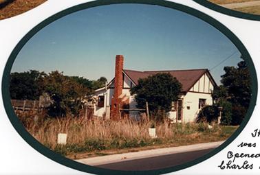 Small house with white/cream painted walls, possibly brick and fibro, red corrugated iron gable roof, with skillion roof on small section at back. External red brick chimney on side wall, wire door at front. There are brown wood straps at intervals on gable ends, and a white trellis fence across back of block. Tall weeds are growing on nature strip.